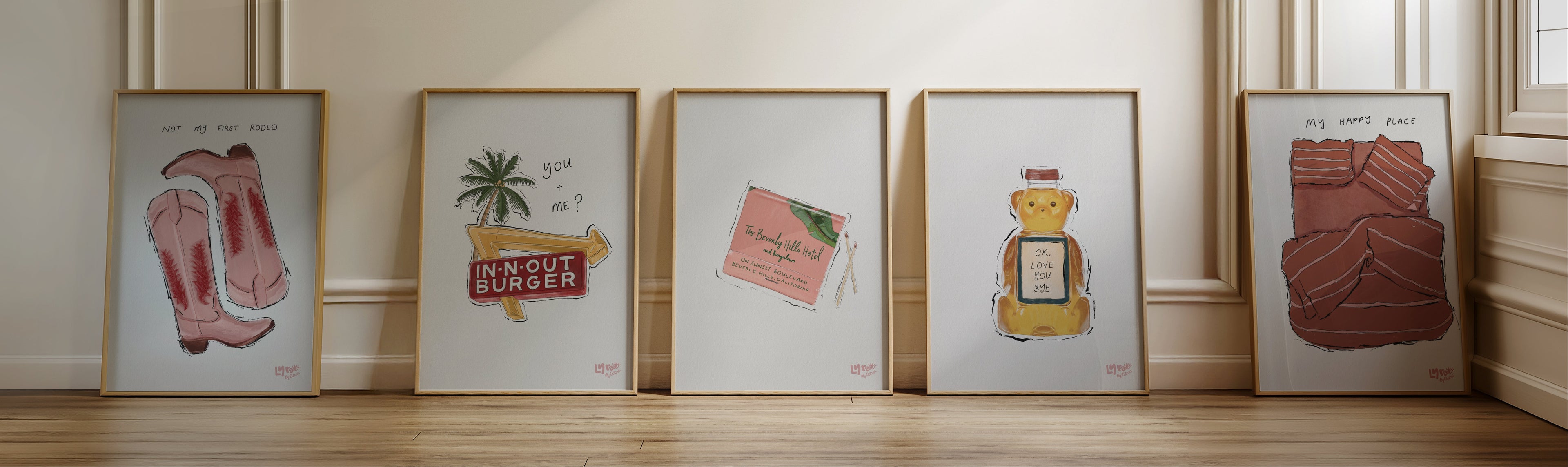 Five framed artworks on a wooden floor against a beige wall.